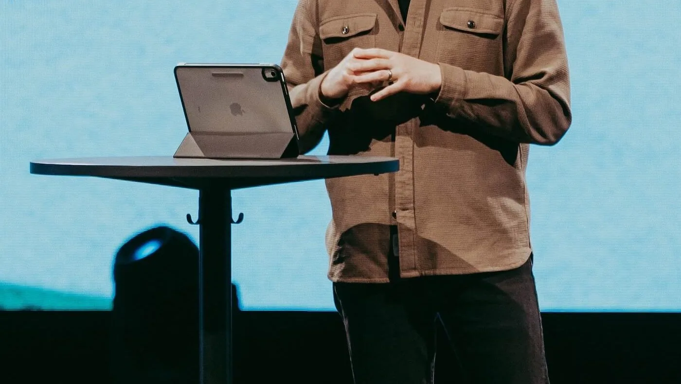 A preacher mid-message, standing beside a small stand with an iPad propped open on it.