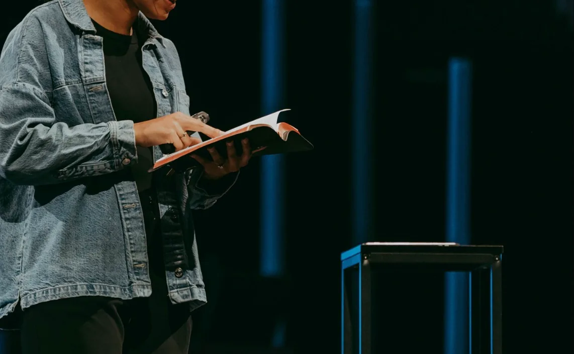 A preacher in a denim jacket holding an open Bible on a dark stage, beside a clear lectern lit in blue.