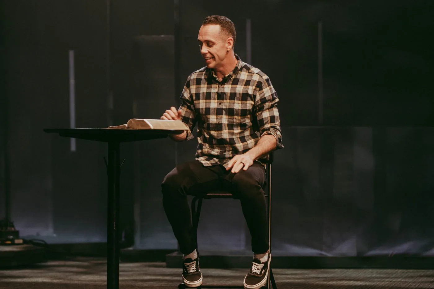 A pastor sitting on a stool beside a stand with an open Bible on it, reading and smiling.