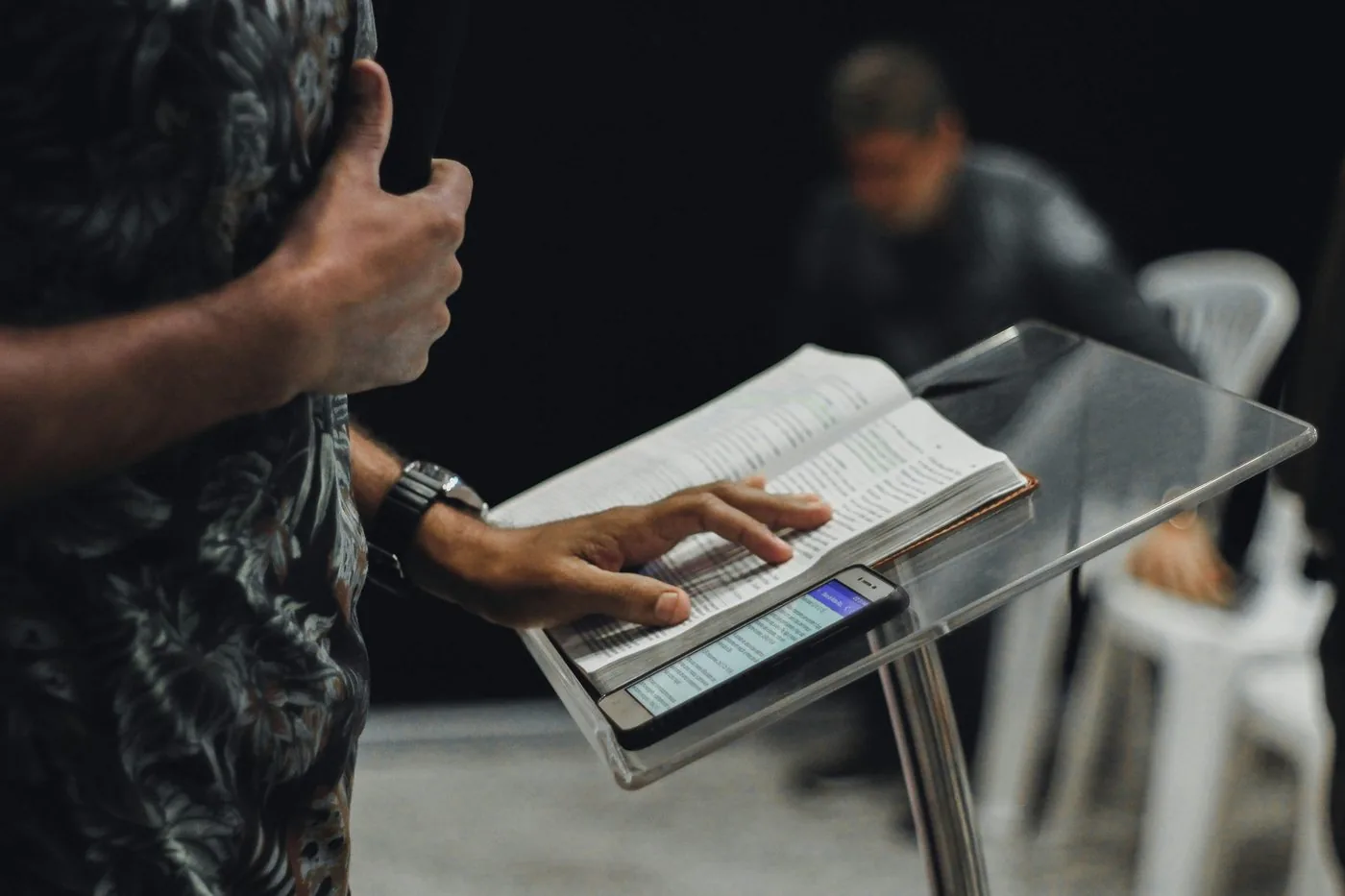 A preacher's hands resting on an open Bible on a clear lectern, with a phone beside it.
